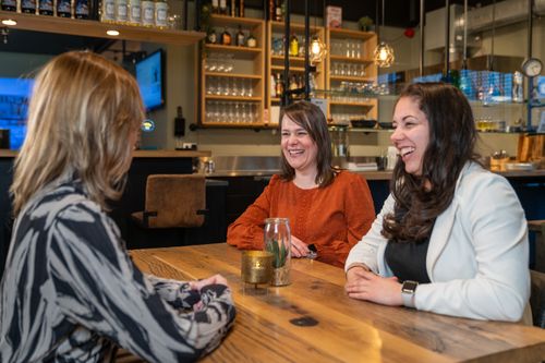 Drie collega's van Rochewood in gesprek en lachen aan een vergadertafel in de lunchruimte van een moderne kantooromgeving