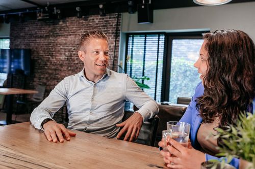 Twee collega's van Rochewood in gesprek aan een vergadertafel met een glas water in de voorgrond