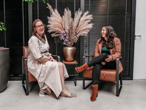 Twee collega's van Rochewood lachen met elkaar zittend in stoelen in een moderne werkomgeving