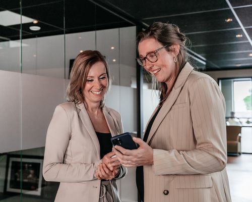 Twee collega’s van Rochewood bekijken samen iets op een telefoon en lachen, in een moderne kantoorruimte