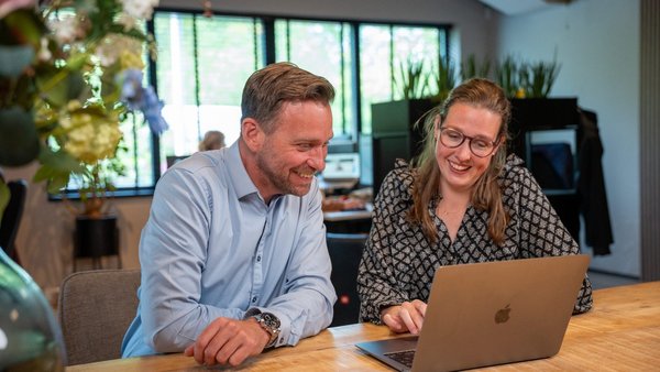 Man en vrouw aan een tafel met een laptop. Vrouw laat wat zien aan mannelijke collega.