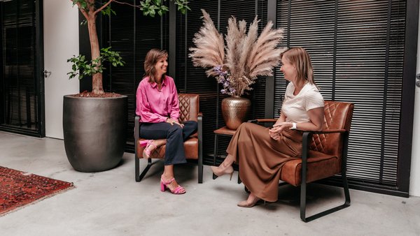 Twee collega's van Rochewood lachen met elkaar zittend in stoelen in een moderne werkomgeving